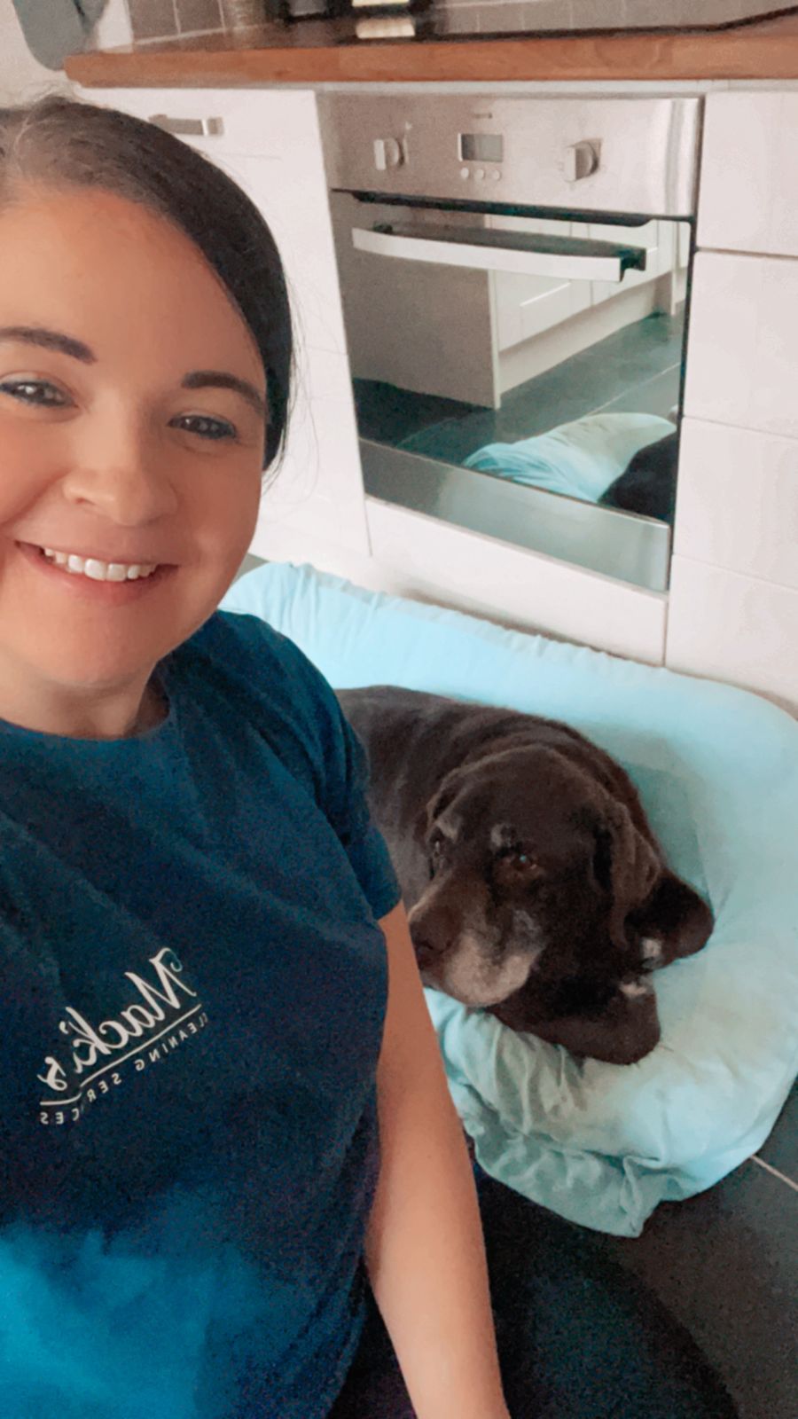 Rhianna at home in a Mackies t-shirt, smiling, with her dog on the sofa behind her